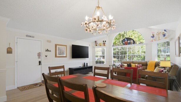 A dining area with a wooden table and chairs, a chandelier, a television on the wall, and a window with a view of greenery. - "castle Retreat" Close To All The Magic! 3 Bedroom Condo (Kissimmee)