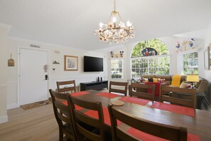 A dining area with a wooden table and chairs, a chandelier, a television on the wall, and a window with a view of greenery. - "Castle Retreat" Close To All The Magic! 3 Bedroom Condo (Kissimmee)