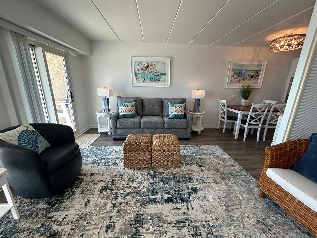 A living room with a grey sofa, a black armchair, a wicker ottoman, a dining table with chairs, and a large rug.