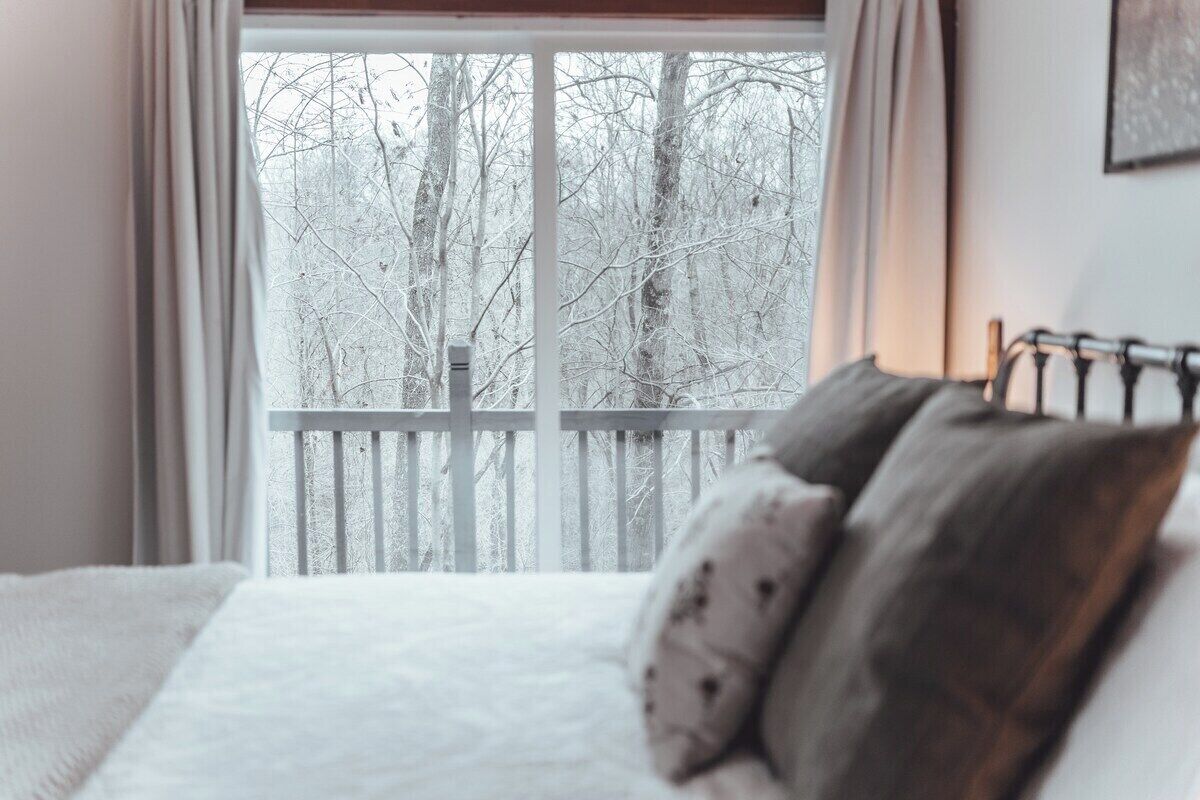 Ein Schlafzimmer mit einem Bett, Kissen und Blick auf einen verschneiten Wald durch ein Fenster.