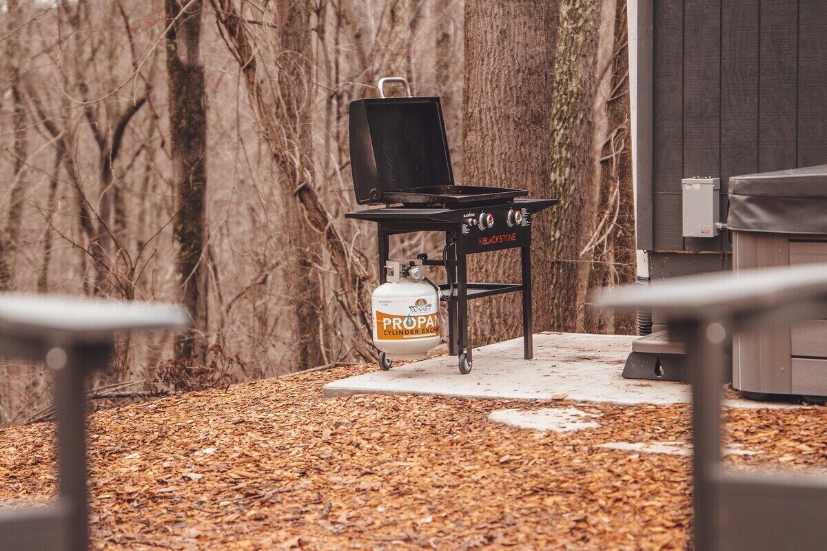 A barbecue grill with a propane tank in a backyard with trees and a building in the background.