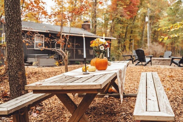 A wooden picnic table with a vase of flowers and a pumpkin centerpiece, set in a wooded area with a cabin in the background.