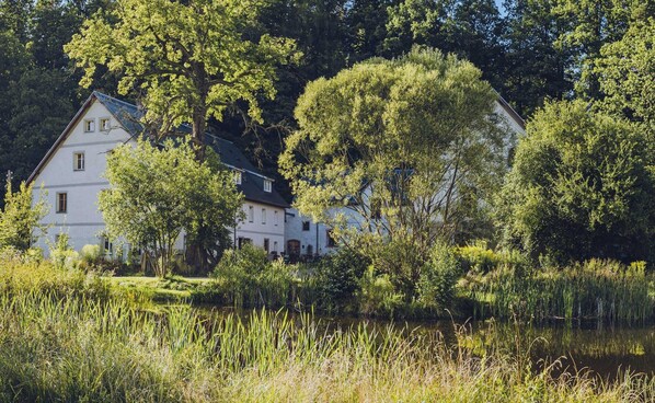 A white building with multiple windows, surrounded by lush greenery and a body of water. - Countryside Stay in Trzcinsko (Janowice Wielkie)