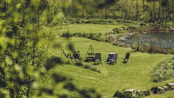 Un espacio al aire libre con césped, un estanque rodeado de árboles y follaje. Varias sillas y una mesa dispuestas para comidas al aire libre.