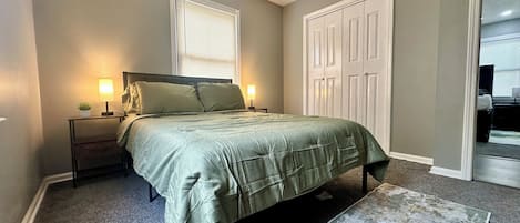A bedroom with a bed, nightstands, a lamp, a window with blinds, and a door.