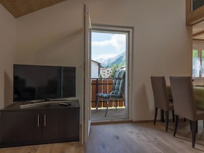 Ein Zimmer mit Fernseher auf einer Kommode, einem Essbereich mit Stühlen und einer Glasschiebetür, die auf einen Balkon mit Bergblick führt.