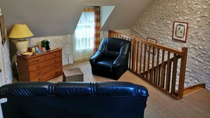 A loft-style room with a sloped ceiling, a wooden staircase, a black leather sofa, a wooden dresser, and a lamp.