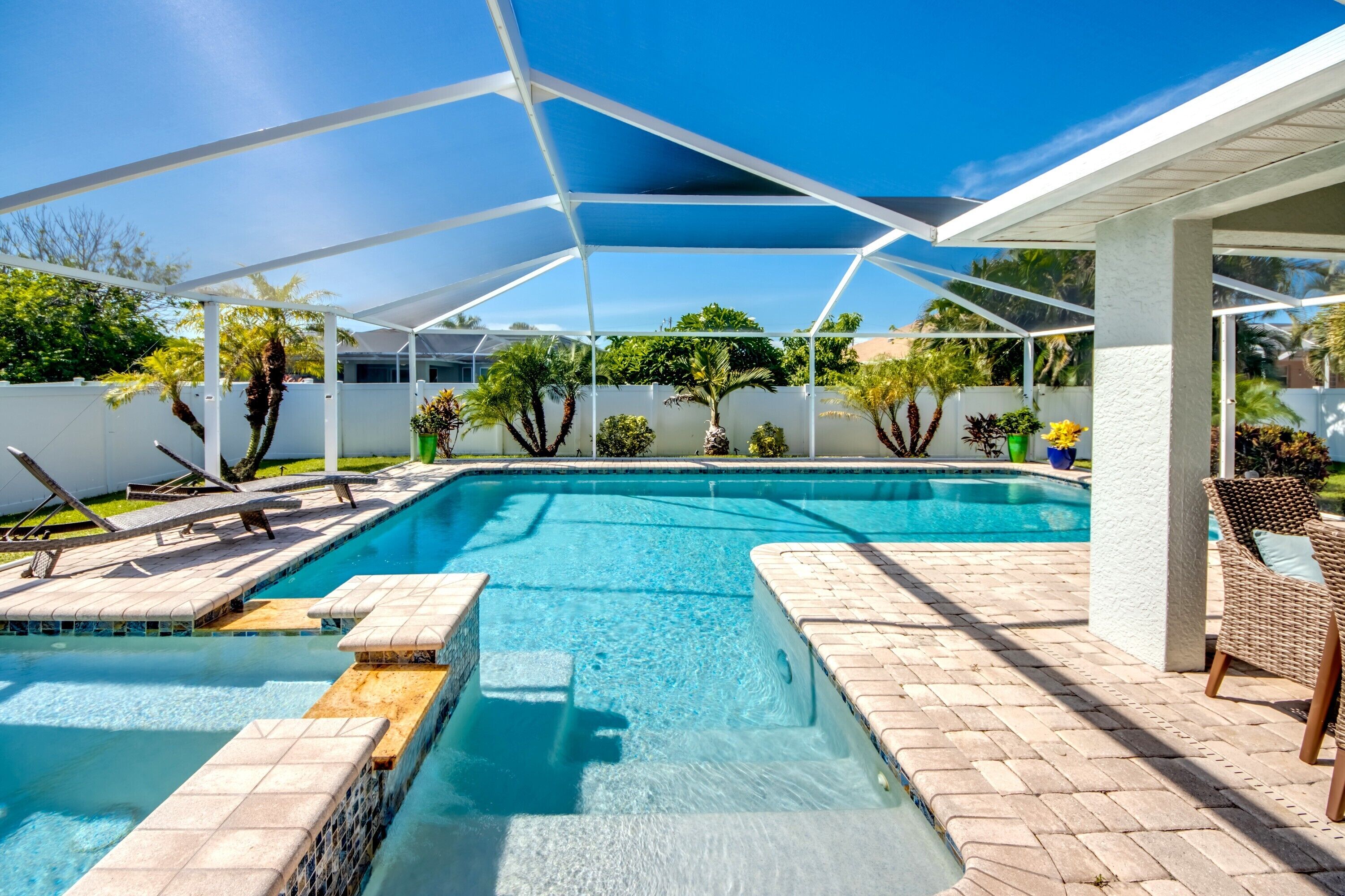 A clear blue swimming pool with a sunshade structure, surrounded by a white fence and various plants.