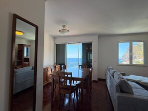 A spacious living area with a dining table, a sofa, and a view of the ocean. - 3-Bedroom Apartment - Camara de Lobos Apartment with Sea and Mountain Views (Camara de Lobos)