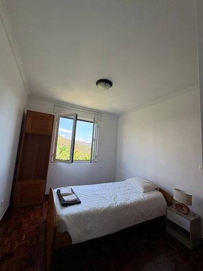 A bedroom with a bed, a nightstand, a lamp, a wardrobe, and a window with a view. - 3-Bedroom Apartment - Camara de Lobos Apartment with Sea and Mountain Views (Camara de Lobos)