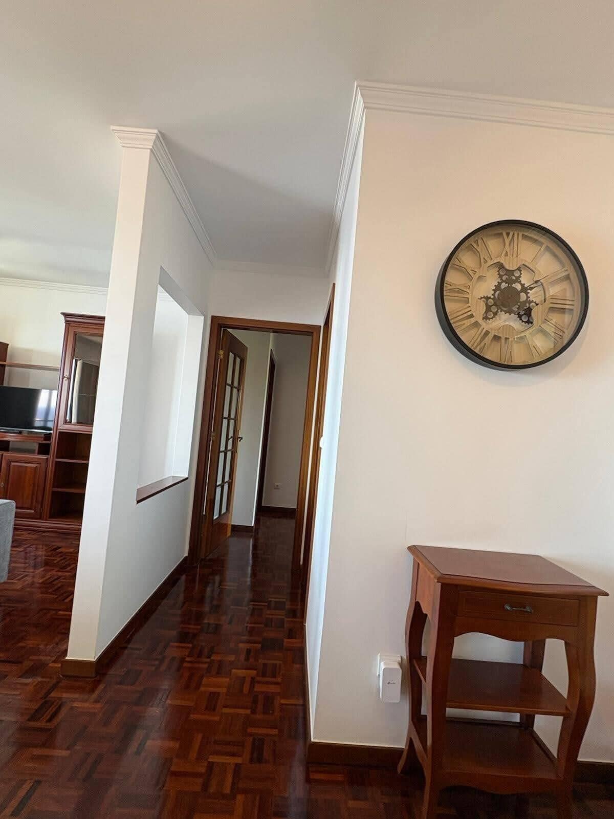 A hallway with wooden flooring, a clock on the wall, and a small wooden table.