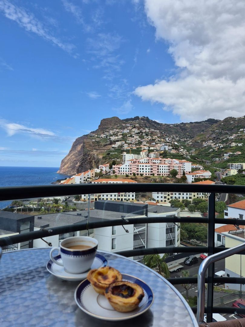 Un balcone con un tavolo, una tazza di caffè e un piatto di cibo, con vista su una cittadina costiera, edifici e scogliere.