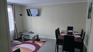 Uma área de refeições com mesa de vidro, cadeiras pretas e uma TV de tela plana fixada na parede.
