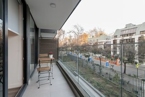 Ein Balkon mit Holztisch und -stühlen, einem Blumentopf und Blick auf die Straße mit Gebäuden und Bäumen.