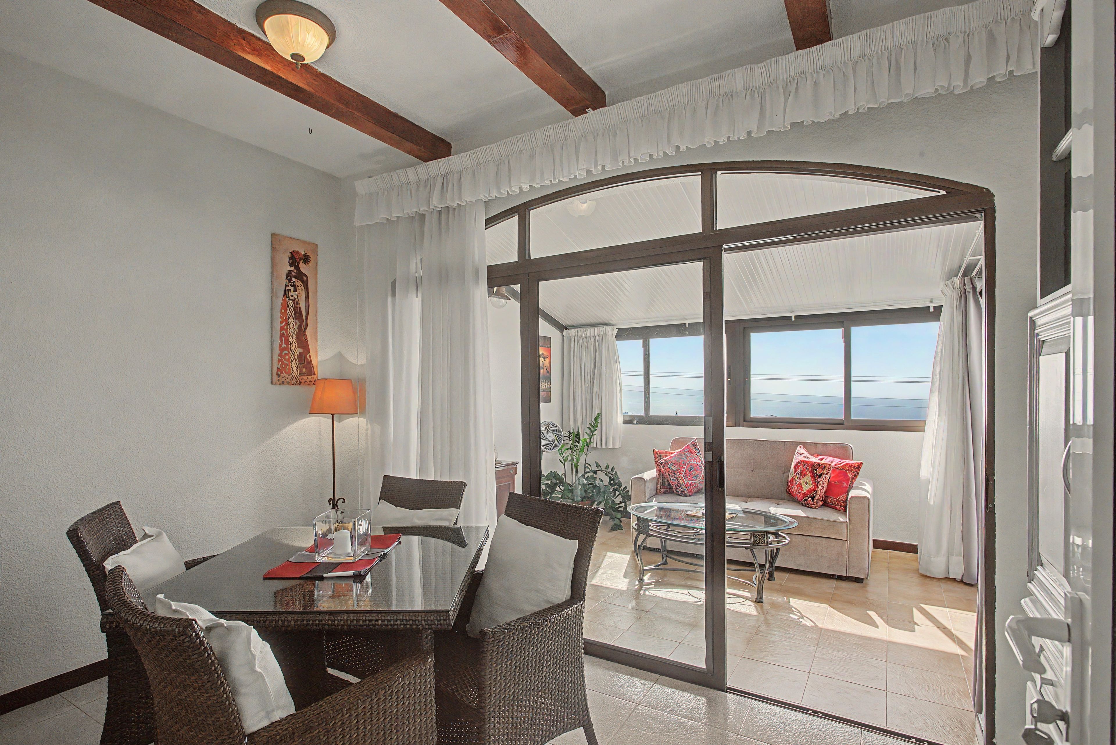 Un coin repas comprenant une table, des chaises et une vue sur un balcon aménagé avec un canapé et des coussins, accessible par une porte vitrée.