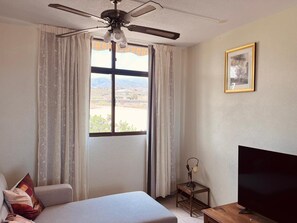 Ein Hotelzimmer mit einem Bett, einem Fernseher, einem Deckenventilator, einem Fenster mit Vorhängen und einem kleinen Tisch.