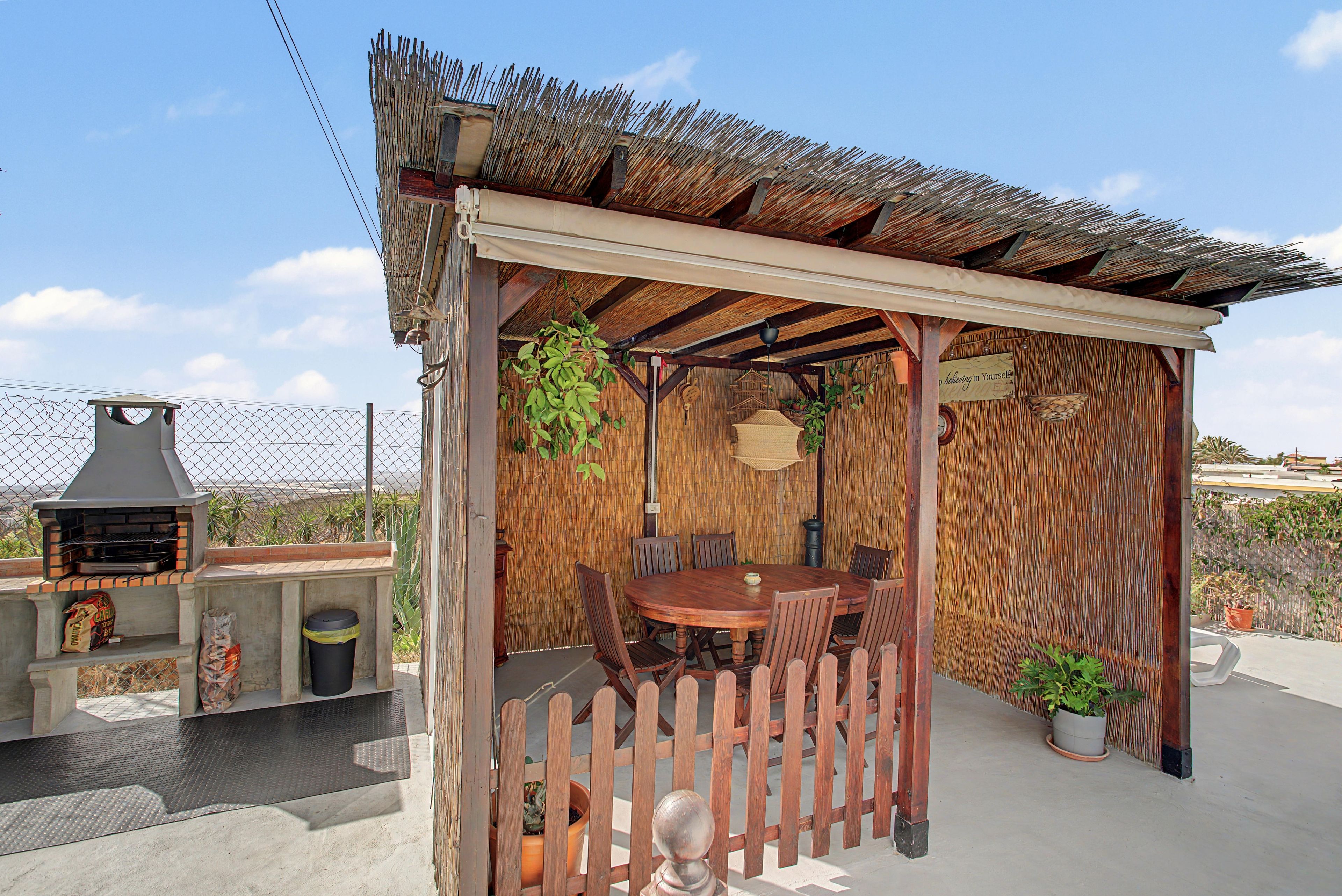 Un patio exterior con mesa y sillas de madera, una parrilla y una pérgola con techo de paja.