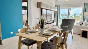 Comedor con una mesa lista para cuatro comensales, sillas con asientos acolchados, una planta en maceta y vistas al exterior.
