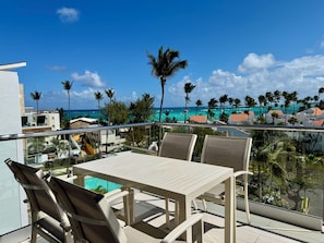 A balcony with a table and chairs overlooking a pool and a coastal view with palm trees. - Gold Coast Luxury Sea View, High-End Amenities (Punta Cana)