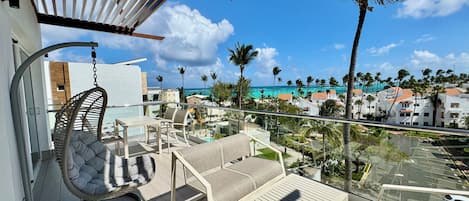 A balcony with a hanging chair, a table, and a sofa, overlooking a coastal area with buildings and palm trees.