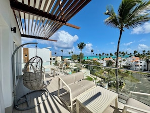 A balcony with a hanging chair, a table, and a view of a resort area with palm trees and buildings. - Gold Coast Luxury Sea View, High-End Amenities (Punta Cana)