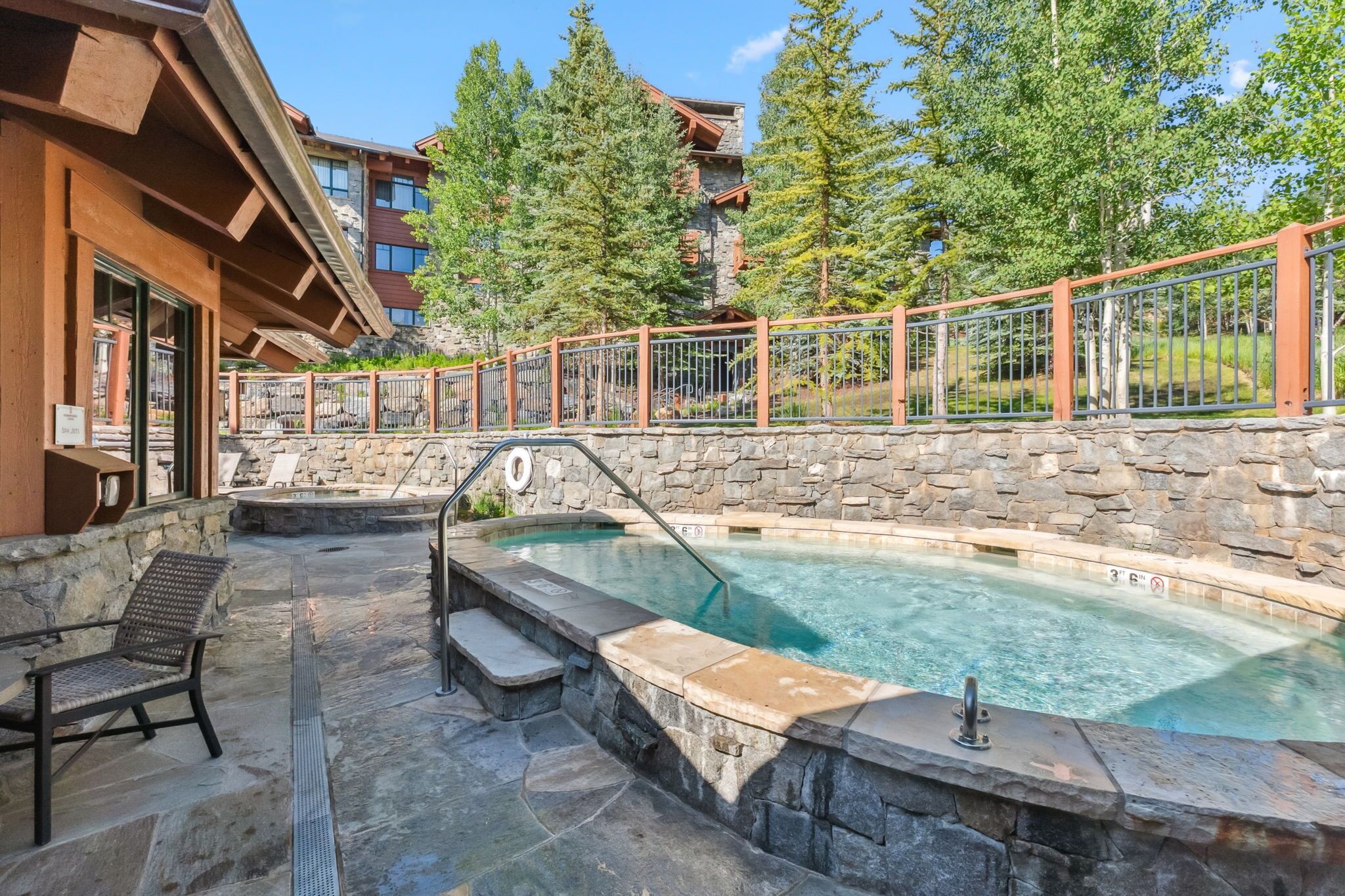 A stone-walled hot tub area with steps leading into the water, surrounded by a wooden deck and a metal fence.