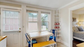 Une pièce avec une porte blanche, une fenêtre avec des stores, un bureau en bois, une chaise bleue, un banc et une étagère contenant des objets rouges et blancs.