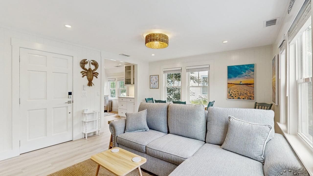 Un salon moderne avec un canapé d’angle gris, une table basse en bois et un coin repas comprenant une table et des chaises blanches.