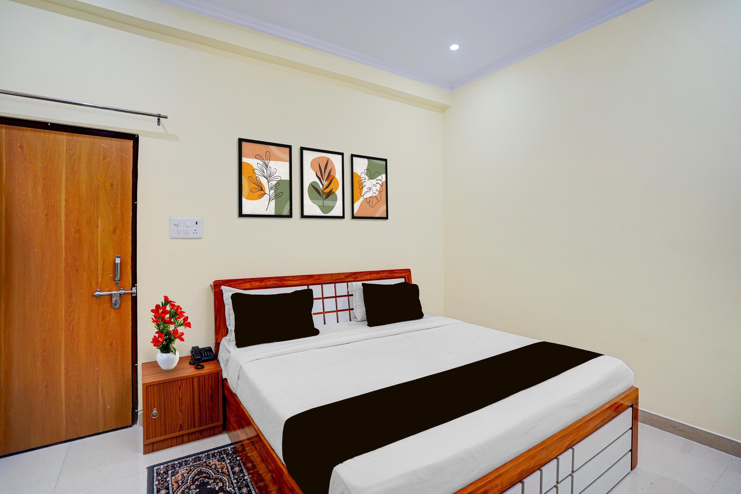 A bedroom with a wooden door, a bedside table with a vase of flowers, a bed with a black and white bedspread, and three framed artworks on the wall.