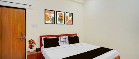 A bedroom with a wooden door, a bedside table with a vase of flowers, a bed with a black and white bedspread, and three framed artworks on the wall.