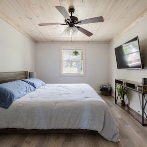 Ein Schlafzimmer mit einem Bett, Deckenventilator, Fernseher, Fenster und einer kleinen Pflanze.