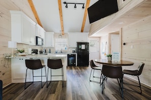 Een moderne keuken met witte kasten, een zwarte koelkast, een eettafel met vier stoelen en een aan de muur bevestigde televisie.
