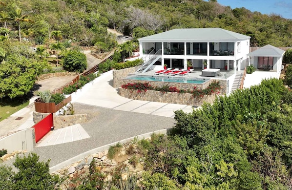 A modern two-story house with a pool, surrounded by lush greenery and a hillside.