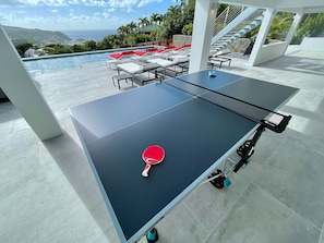A ping pong table with a paddle, set up outdoors with a pool and lounge chairs in the background.