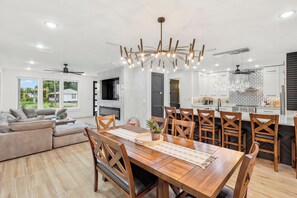 A modern kitchen with a central island, overhead lighting, and a dining area with a wooden table and chairs. - Luxury Coastal Retreat | Pool, Golf Cart & Gulf Views (Panama City Beach)