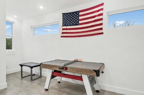 A room with a foosball table, an American flag, and two windows with views of the sky and trees. - Luxury Coastal Retreat | Pool, Golf Cart & Gulf Views (Panama City Beach)