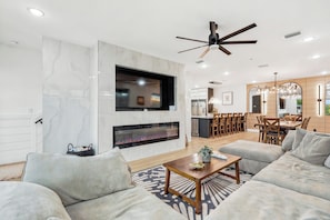 A modern living room with a fireplace, a ceiling fan, and a dining area with wooden furniture. - Luxury Coastal Retreat | Pool, Golf Cart & Gulf Views (Panama City Beach)