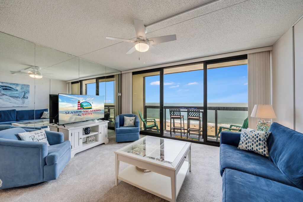 Una sala de estar moderna con televisor, sofás, una mesa de centro y un balcón con vista a la playa.