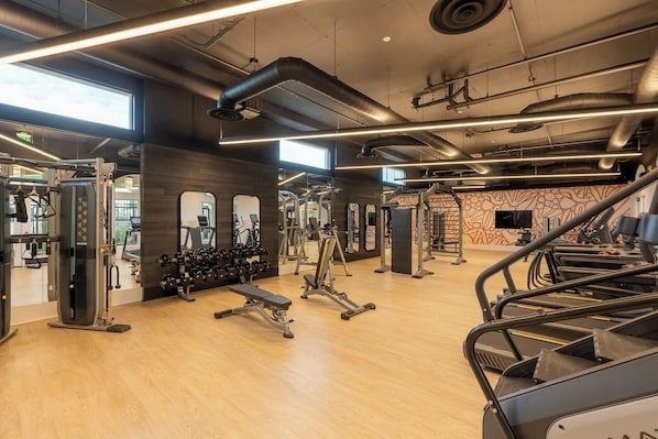 A gym with various exercise equipment, wooden flooring, and large windows allowing natural light.