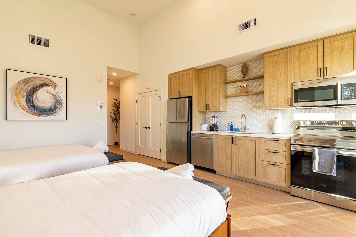 A modern hotel room with two beds, a kitchenette with stainless steel appliances, and a herringbone-patterned backsplash.