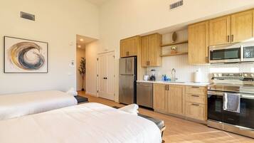 A modern hotel room with two beds, a kitchenette with stainless steel appliances, and a herringbone-patterned backsplash.