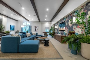 Una sala de estar moderna con un sofá seccional, una mesa de centro y un televisor montado en la pared. Hay plantas en macetas y un mural floral grande en la pared.