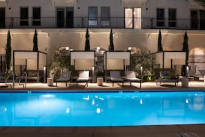 Una piscina con sillas de descanso y sombrillas, rodeada por un edificio con balcones y una valla.