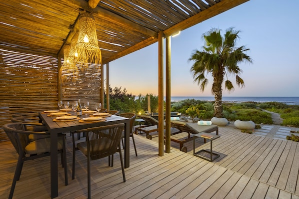 An outdoor dining area with a wooden deck, tables set for dining, and a view of the sea.