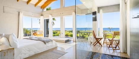 A modern bedroom with a large bed, a view of the sea, and a glass-enclosed bathroom with a bathtub.