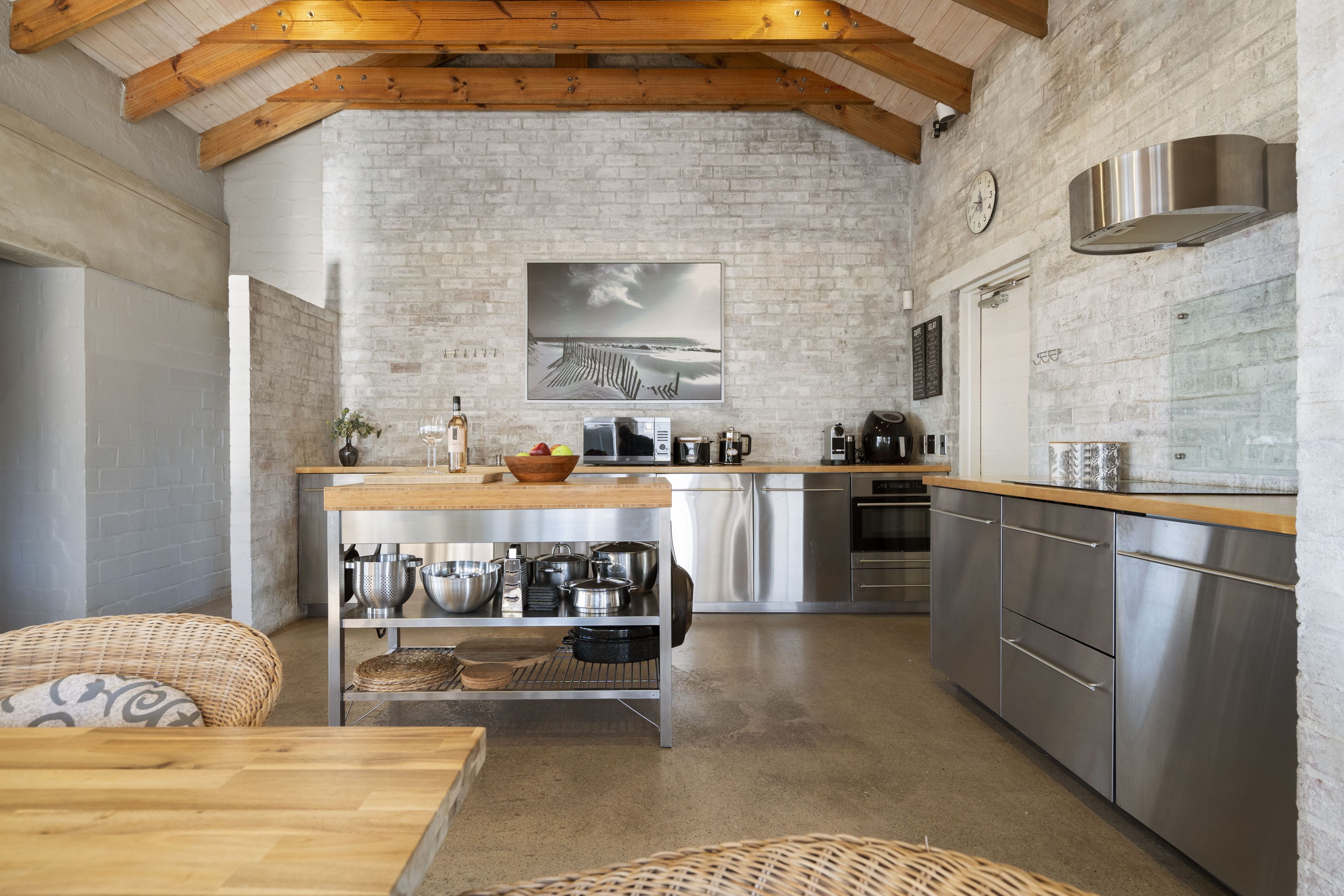 A compact kitchen with a brick wall, wooden beams, stainless steel appliances, and a dining area with wicker furniture.