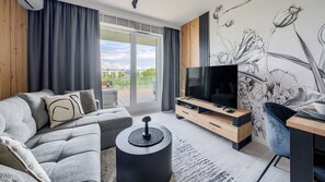 Una sala de estar moderna con un sofá gris, un televisor de pantalla plana, un mueble de televisión de madera y un ventanal con vistas a zonas verdes.