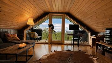 A cozy attic room with a sloped ceiling, a large window, a leather rug, a desk with a chair, and a sofa.