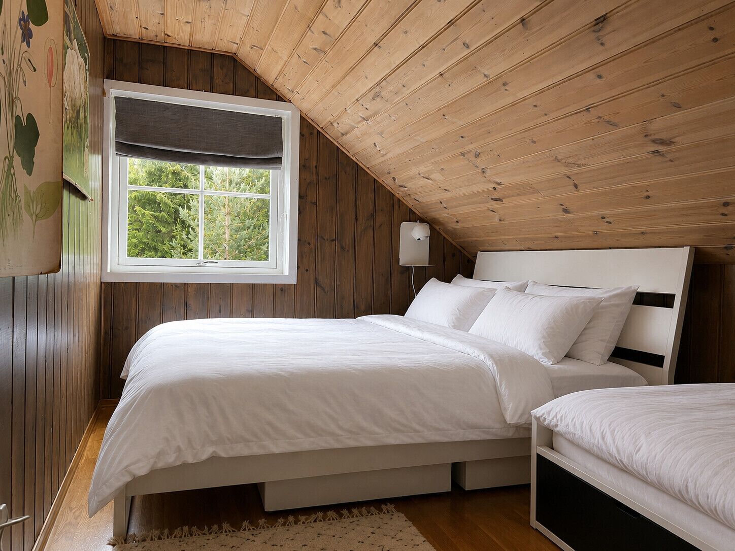 Een slaapkamer met een schuine plafond, een raam met uitzicht op groen, een bedlampje en een bed met wit beddengoed.