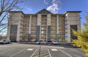 Ein mehrstöckiges Hotel mit zentralem Eingang, Parkplatz und mehreren parkenden Fahrzeugen davor.